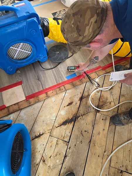 Worker using a handheld moisture meter on flooring beside a marked test location.