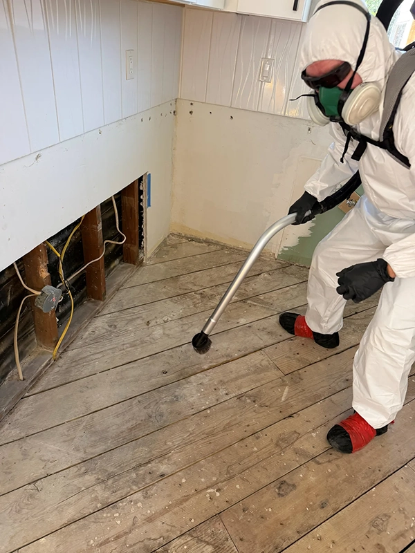 Worker in protective coveralls vacuuming exposed wood flooring beside an opened wall.
