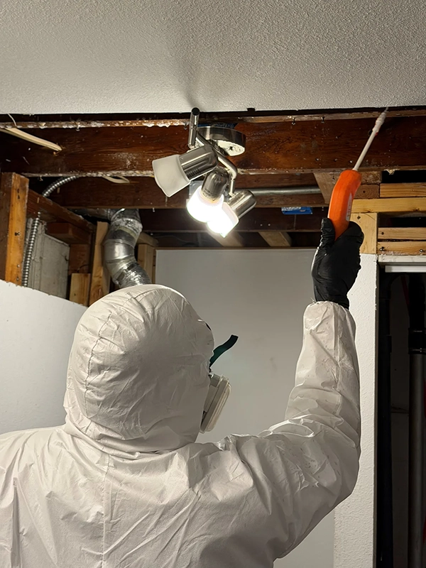 Worker in protective coveralls reaching into an opened ceiling with a hand tool.