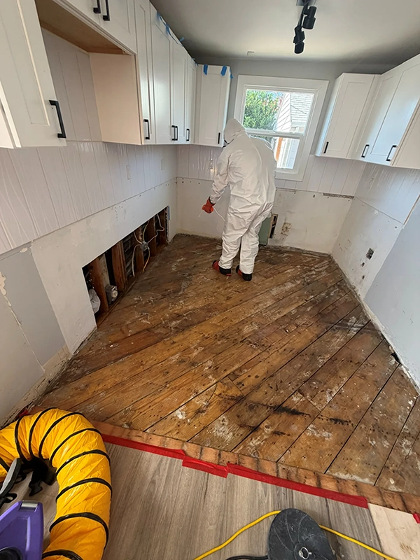 Wide view of a kitchen with exposed wood flooring, opened lower walls and a worker in protective coveralls.
